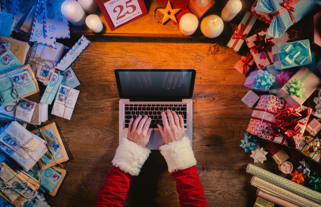 Holiday employee appreciation ideas: Santa typing on a laptop surrounded by gifts on desk